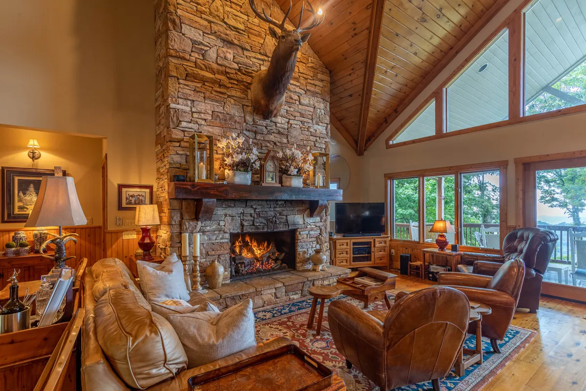 This image features a grand living room showcasing a large stone fireplace set beneath soaring ceilings. The space is adorned with comfortable seating and a mounted deer head above the mantle. Expansive windows bring in natural light and offer scenic views, while the arrangement is ideal for both cozy relaxation and lively social gatherings.