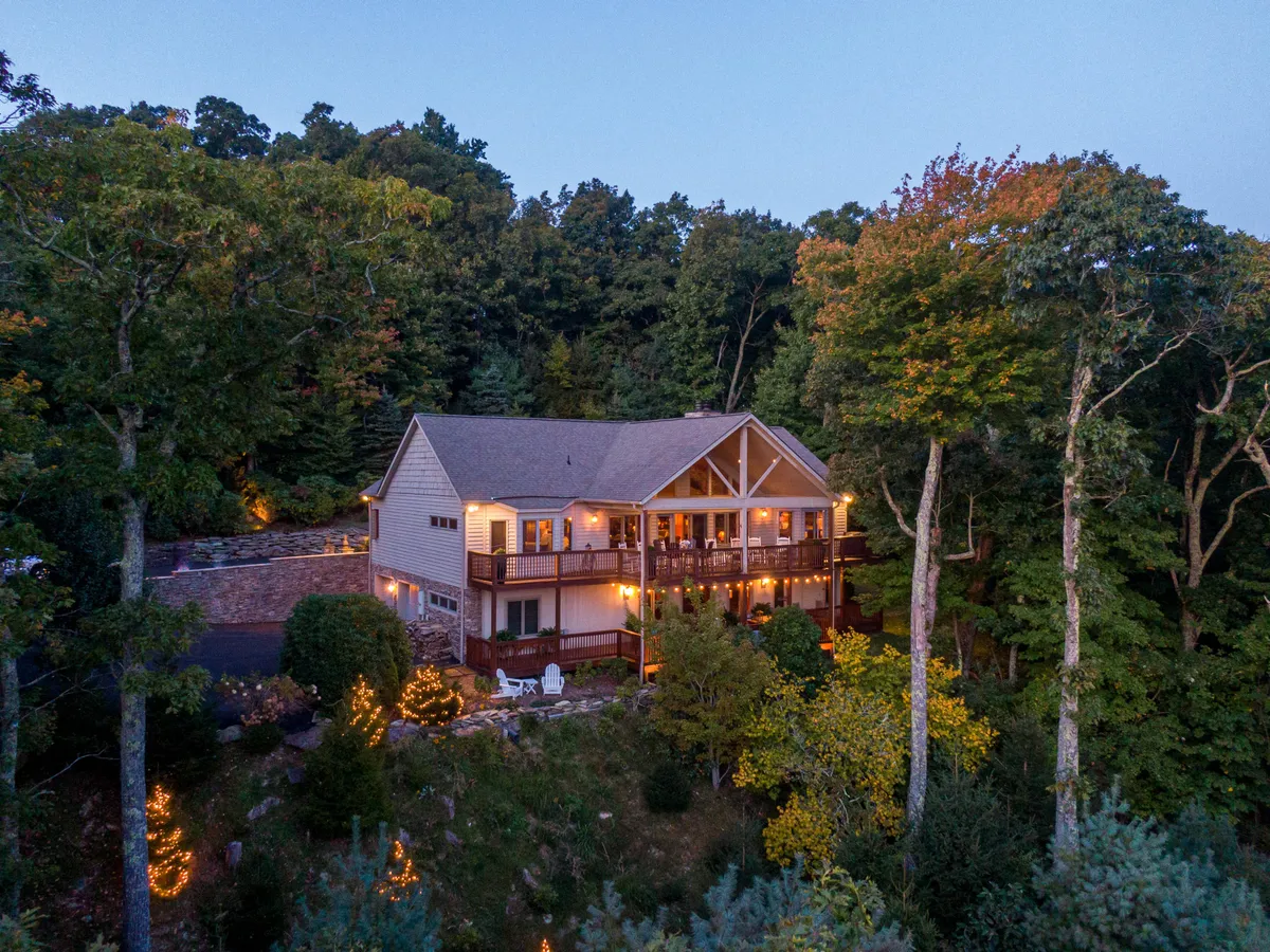 The image showcases a beautiful, spacious mountain home surrounded by towering trees, with a warmly lit deck perfect for evening gatherings. The expansive deck features inviting lighting, offering sweeping long-range views and a welcoming space for guests to relax as the sun sets over the Blue Ridge Mountains.