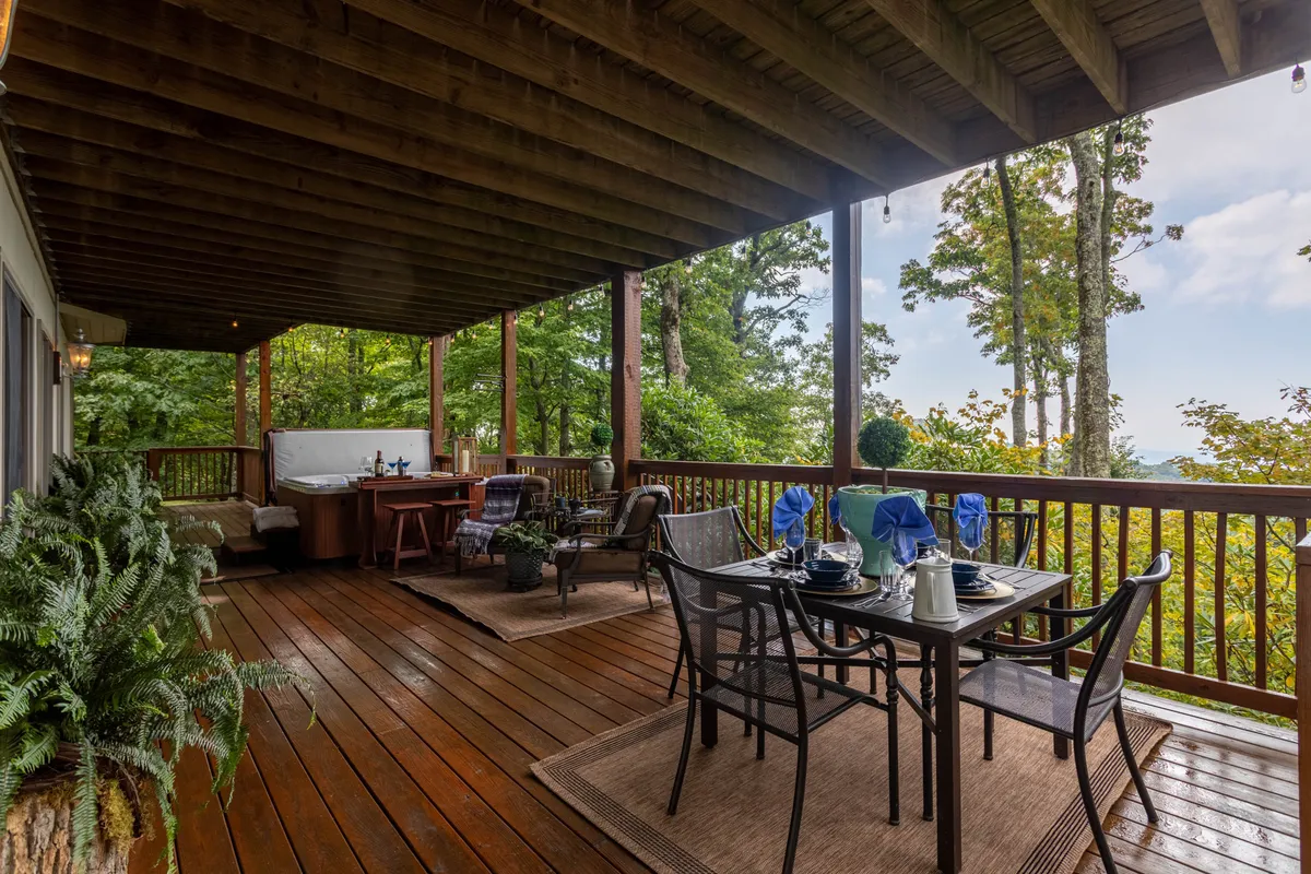A relaxing outdoor setup featuring a dining table with set place settings, a hot tub, surrounding greenery, and a wooden railing. The scene feels like an elevated mountain retreat, ideal for gathering with friends and family while enjoying fresh air and nature.