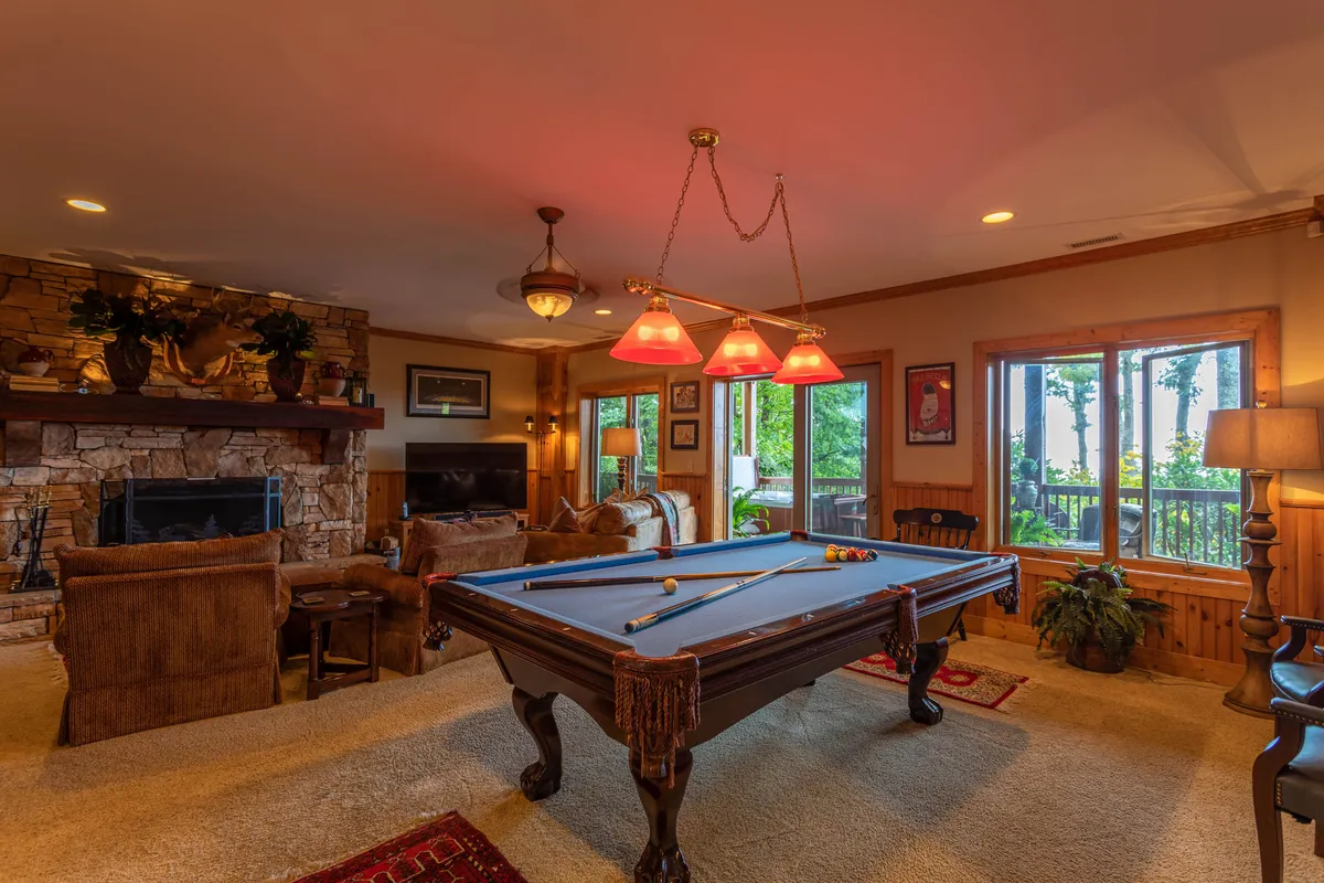 A spacious game room featuring a pool table at the center, a stone fireplace with cozy seating nearby, large windows letting in natural light, and elegant chandeliers overhead. The atmosphere is both relaxed and social, inviting guests to unwind, play, and gather.