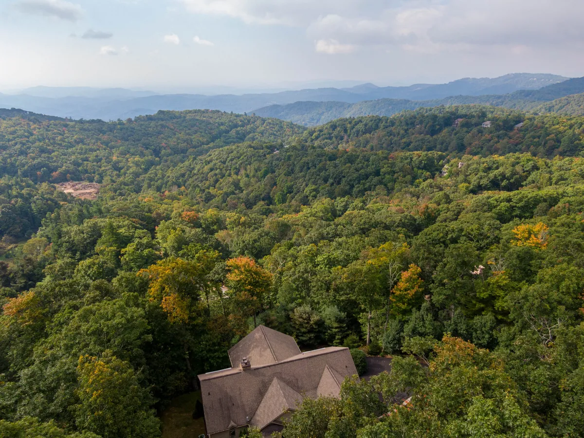 This image captures a sweeping view of a mountain range with a dense forest below and a house nestled in the foreground. The expansive sky and undisturbed natural landscape evoke a sense of tranquility and connection to the surrounding wilderness.