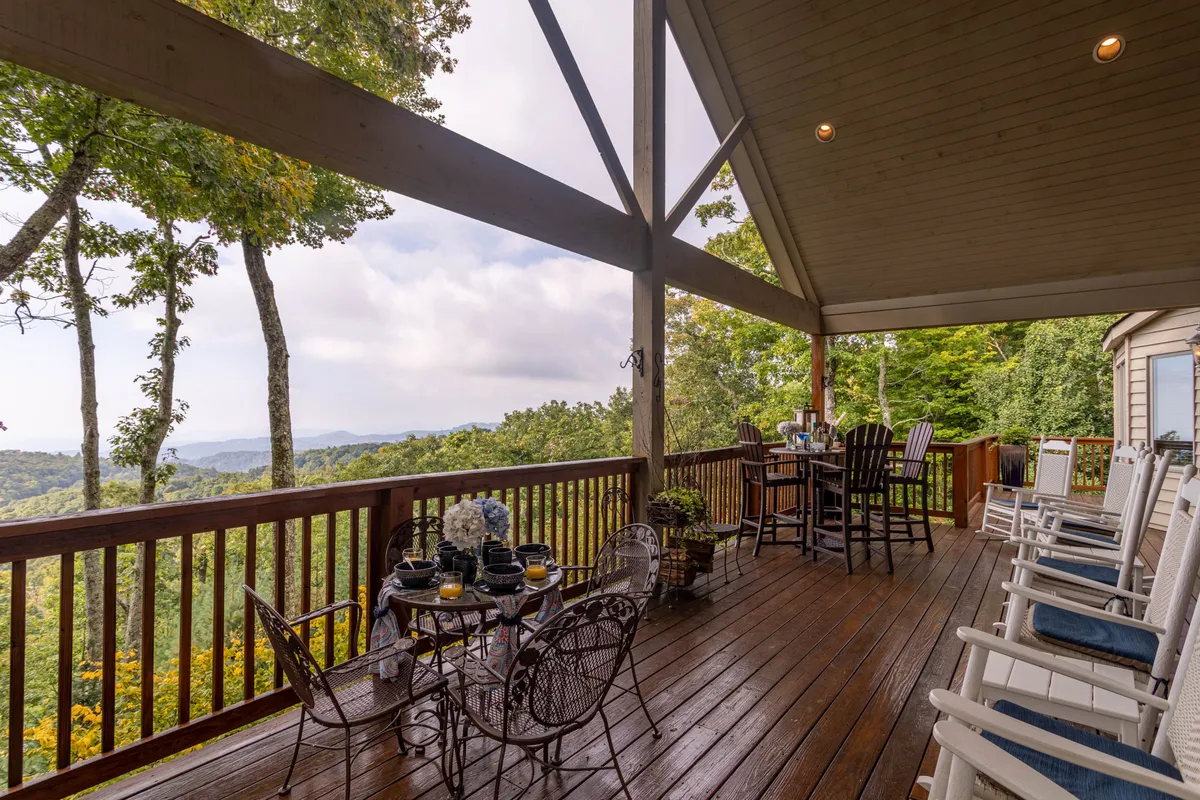 A serene outdoor deck furnished with rocking chairs, a dining table, and bar stools, all set against a stunning mountain view. The setup is ideal for enjoying morning coffee, outdoor dining, or simply soaking in the peaceful atmosphere.