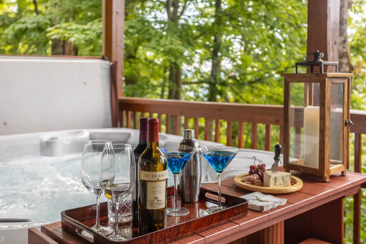 An outdoor hot tub scene with wine and martini glasses on a wooden tray, a grapes and cheese platter, and a glowing lantern. The bubbling hot tub is surrounded by peaceful natural scenery, creating a sense of relaxation and luxury.