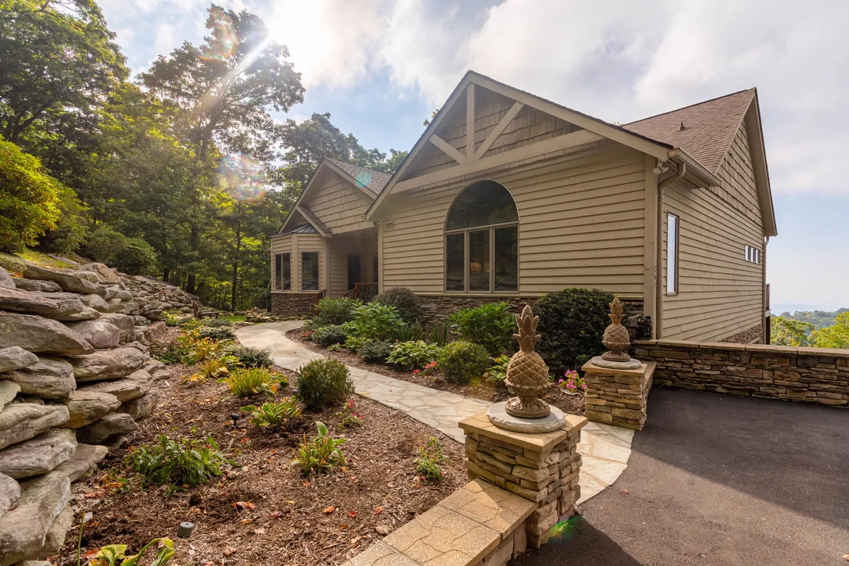 A beautifully landscaped front garden with a stone pathway leading to the house, lined with greenery and decorative statues. Large windows offer glimpses inside, and a serene porch area provides a welcoming first impression.