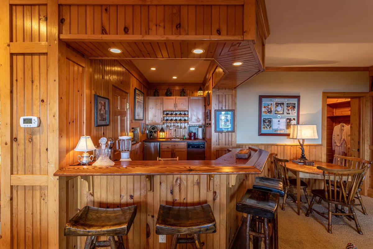 A warm, rustic wet bar area with a wooden bar counter, comfortable seating, and decorative lighting. The bar is set against a backdrop of kitchen amenities and is designed for gathering and entertaining.